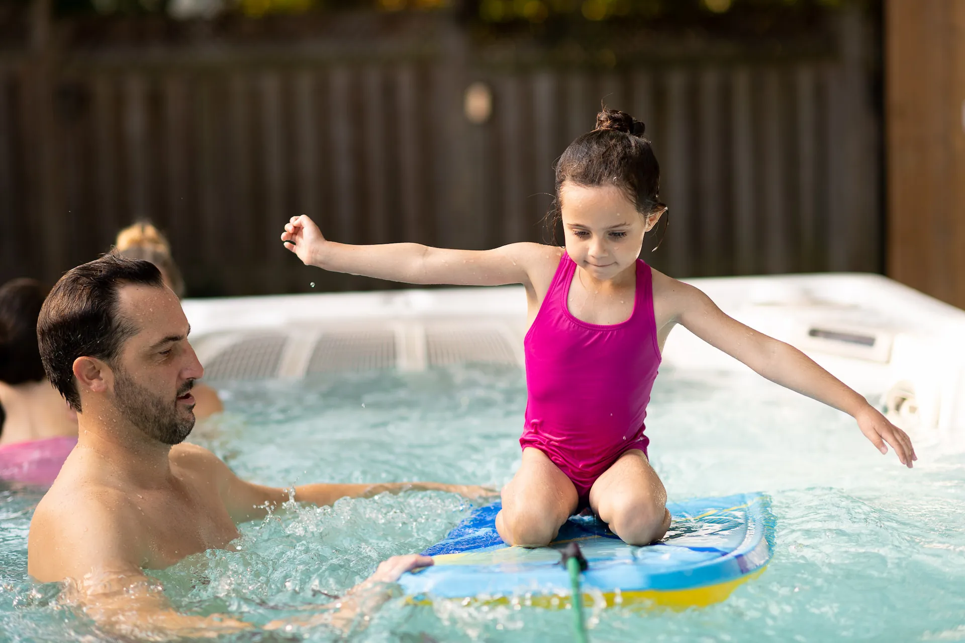 Close up of an adult holding up a child who is kneeling on a boogie board inside a swim spa.