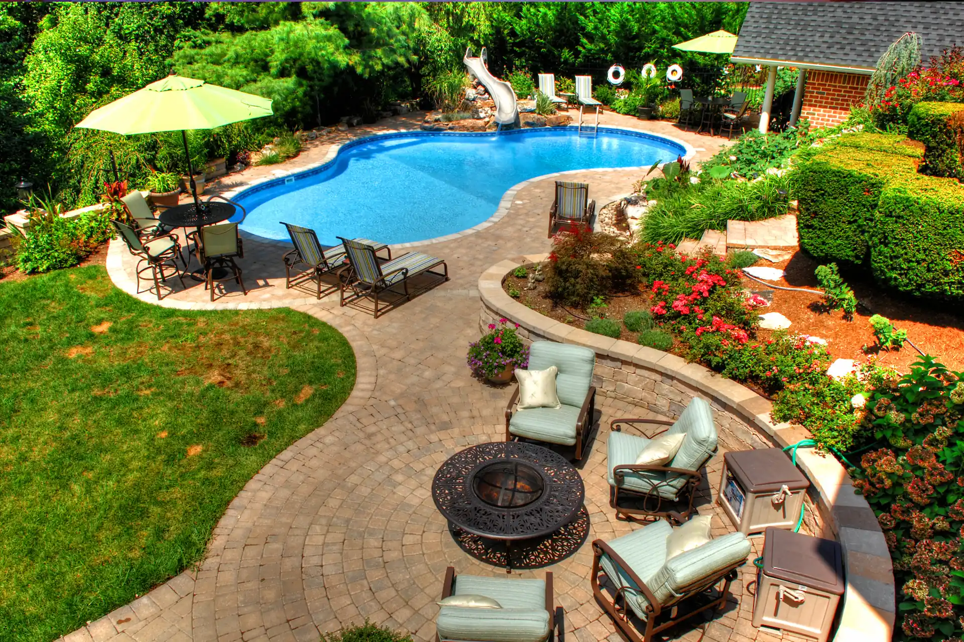 Aerial shot of a backyard with an inground pool surrounded by a bricked pool deck in the daylight. Backyard designs in Central Pennsylvania can include swimming pools and fire pits like those pictured here.