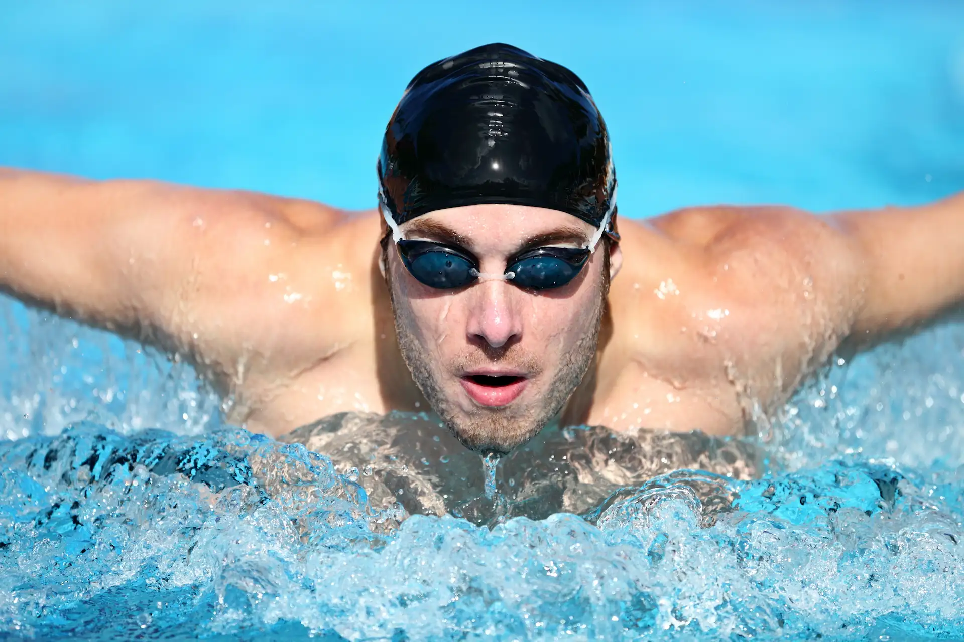 Close-up of a swimmer in dark goggles and a black swim cap performing the butterfly stroke. Activities like these figure into your decision of whether to get a pool or a swim spa.